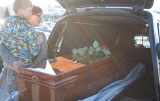 Nainen katselee ruumisarkkua auton takakontissa ja pitelee sylissään nuorta poikaa.