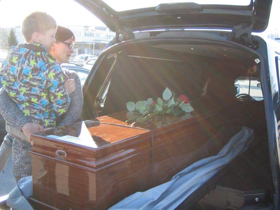 Nainen katselee ruumisarkkua auton takakontissa ja pitelee sylissään nuorta poikaa.